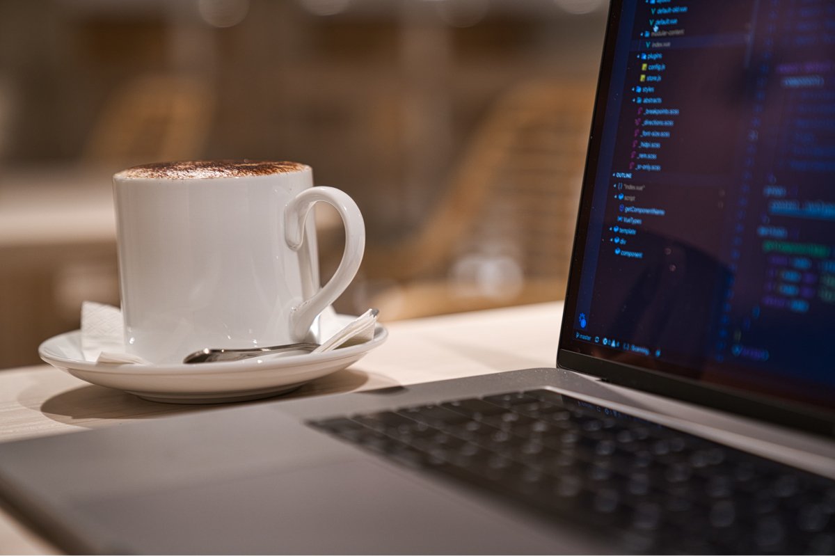 A photo of white coffee mug on a saucer next to a laptop on a cafe table.