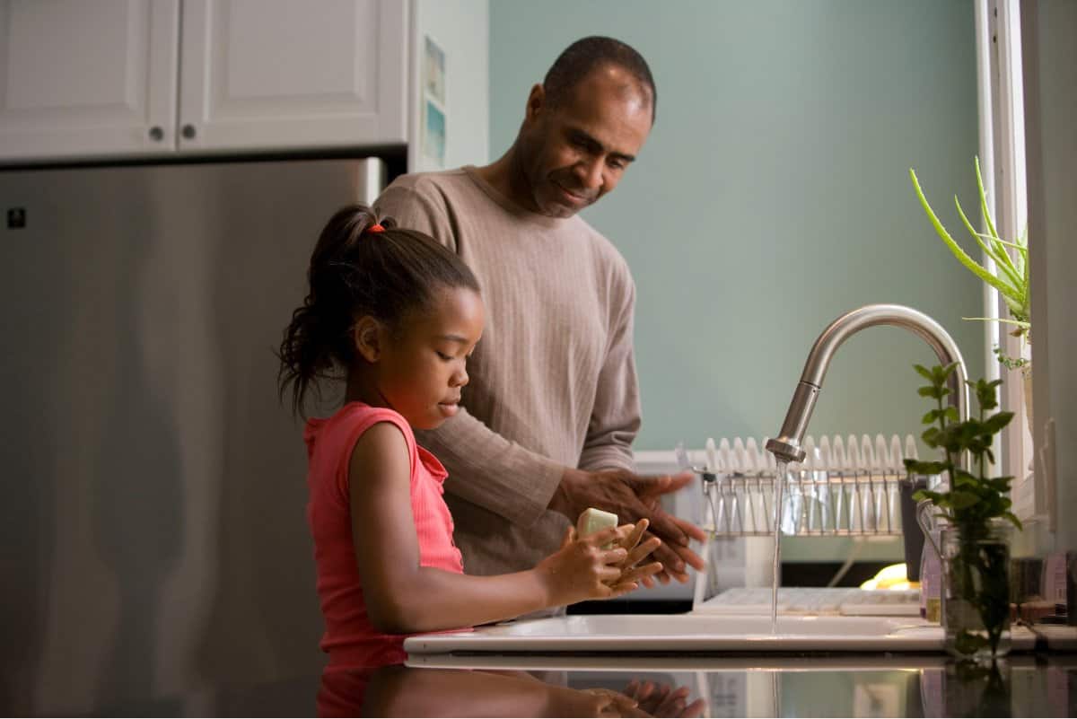 A photo of a father and daughter washing their hands together at the kitchen sink.