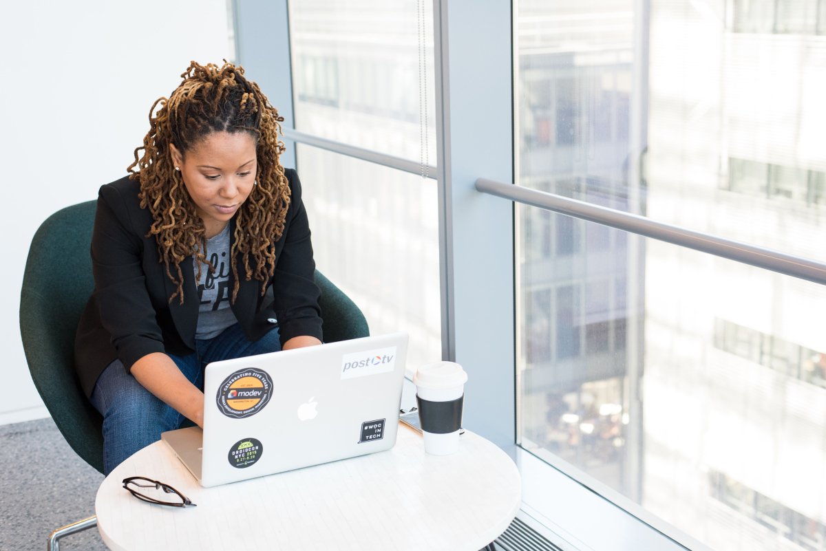 A photo of a woman working on her laptop next to large windows.