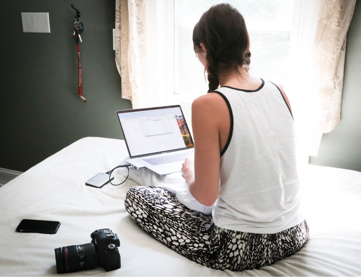 A photo of a woman sitting on her bed with a laptop propped up on a pillow in front of her.