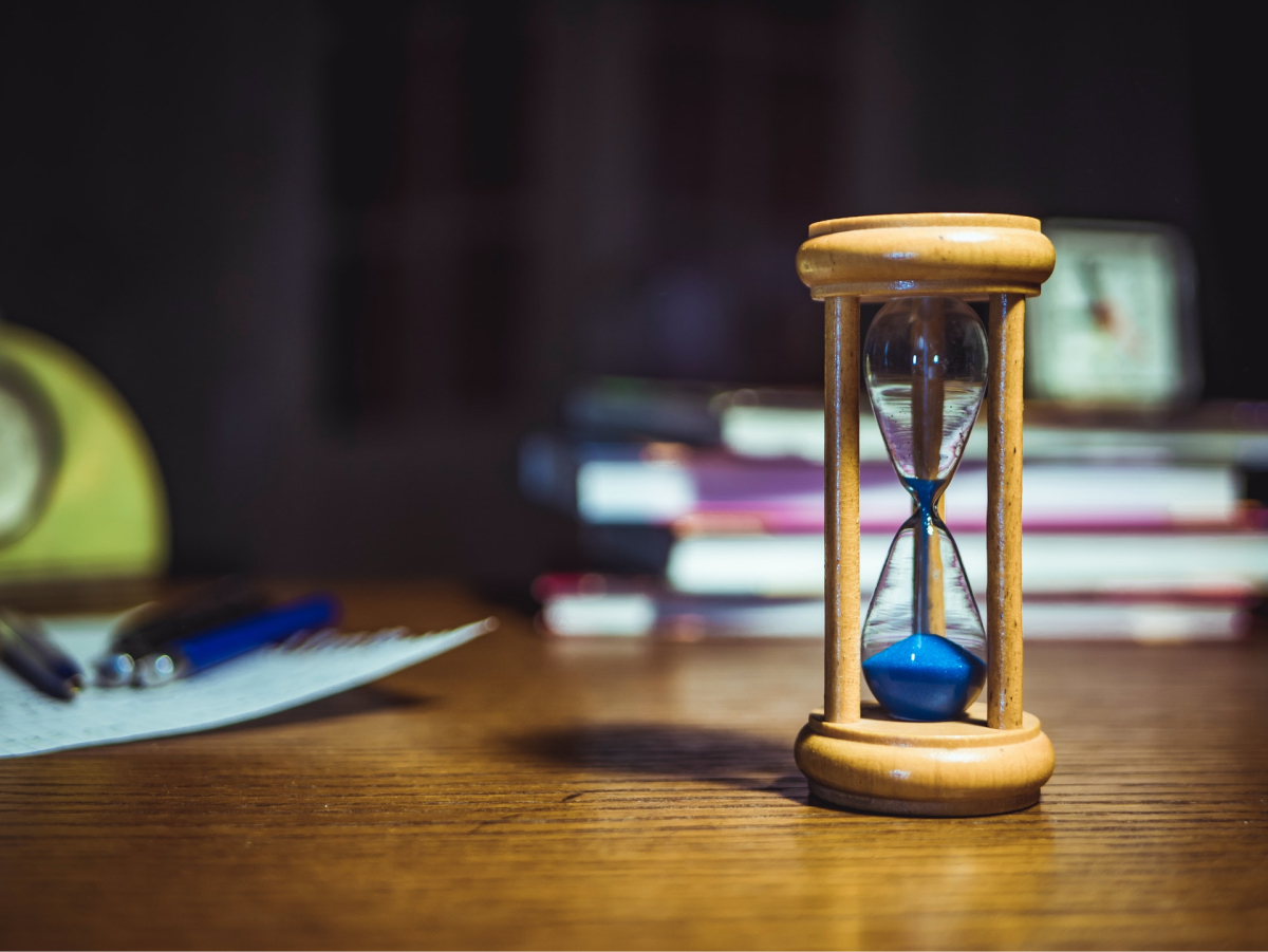  An hourglass sitting on a desk with books and pens in the background. 