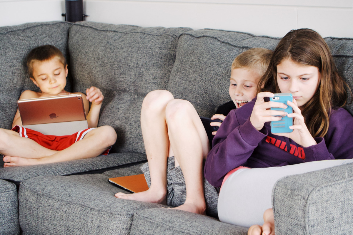 Three children sitting on a couch, each enthralled with a mobile device. 