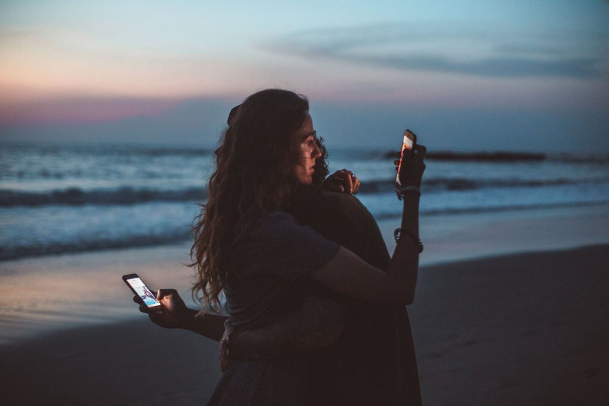 Two people embracing on a beach while looking at their smartphones. 