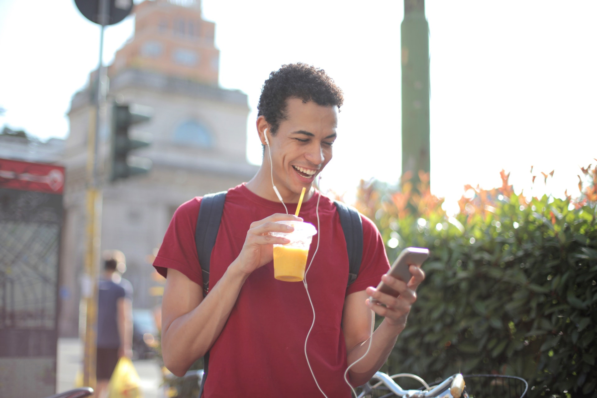A smiling man walking down the street drinking juice and using his phone.
