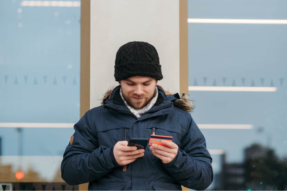  A man in winter clothes entering his credit card number into his smartphone.