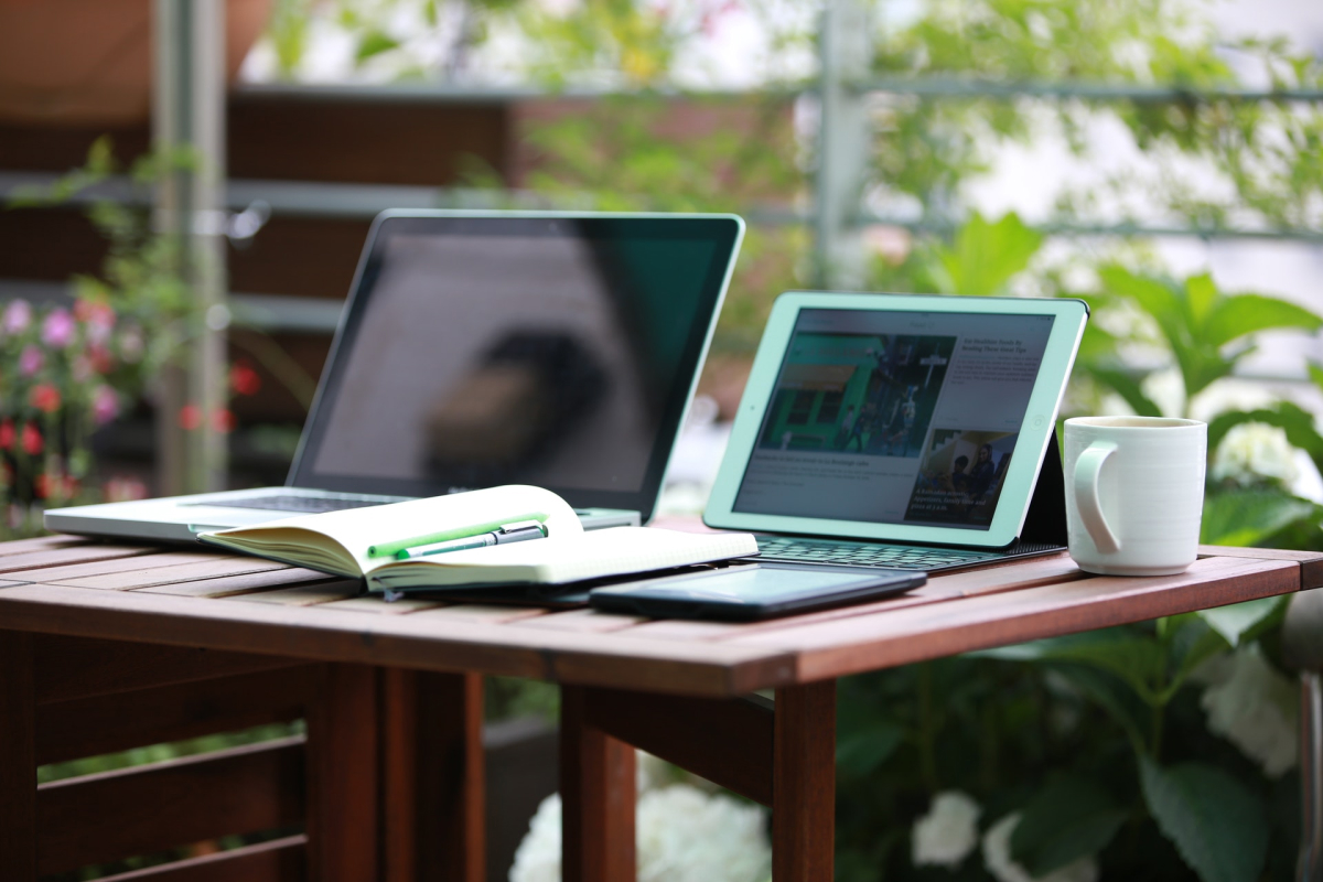 Two laptops sitting on a wooden table with a notebook and mug. 